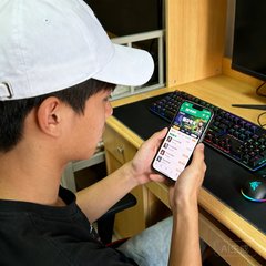 23岁男性电竞爱好者李明轩的生活照，戴着白色棒球帽，手持手机正在查看蜂鸟电竞APP赛事页面，背景为大学宿舍书桌，桌面上摆放着机械键盘和电竞鼠标。